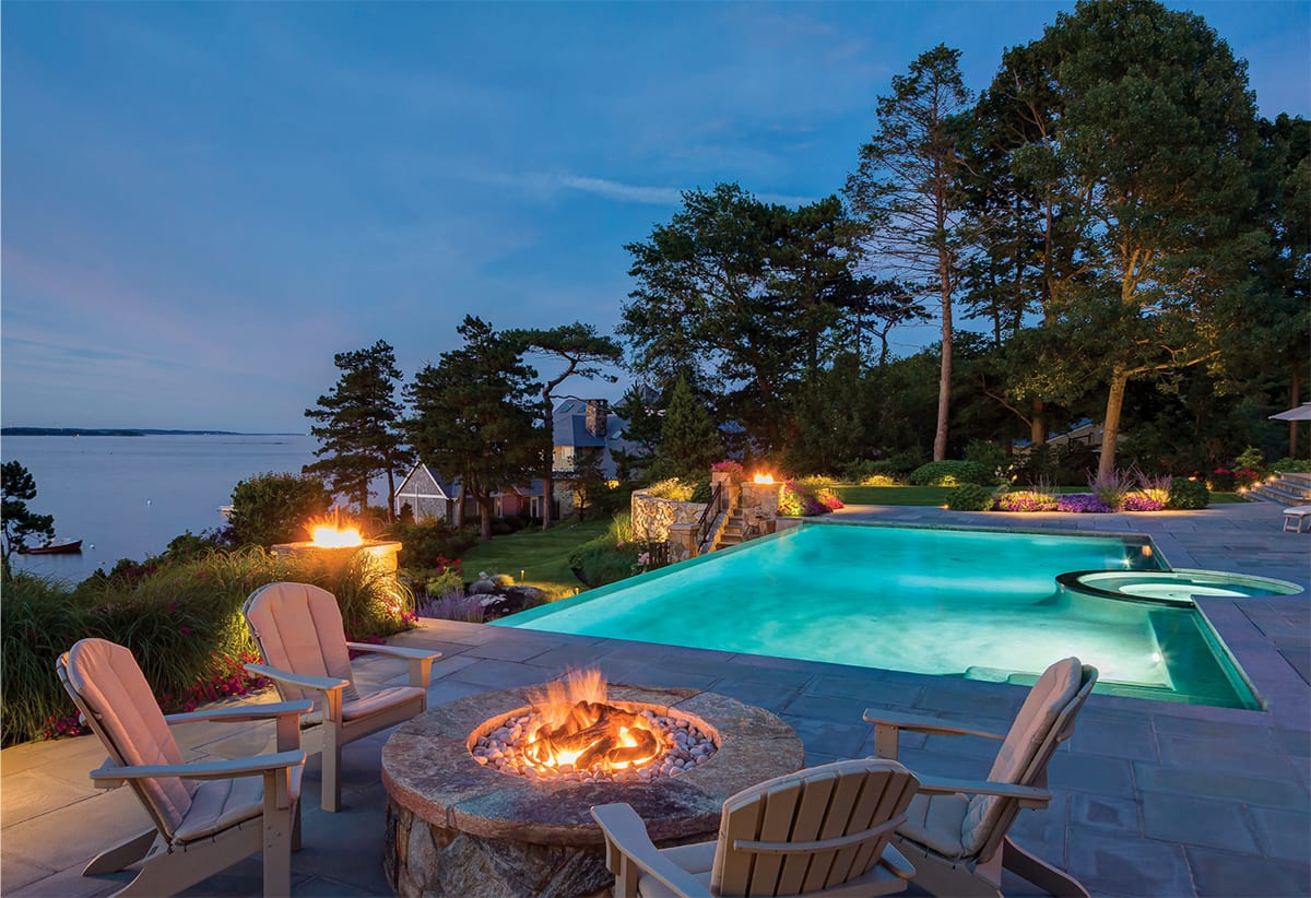 Picture of a pool and hot tub in the evening surrounded by fire pits and with a view of the ocean.