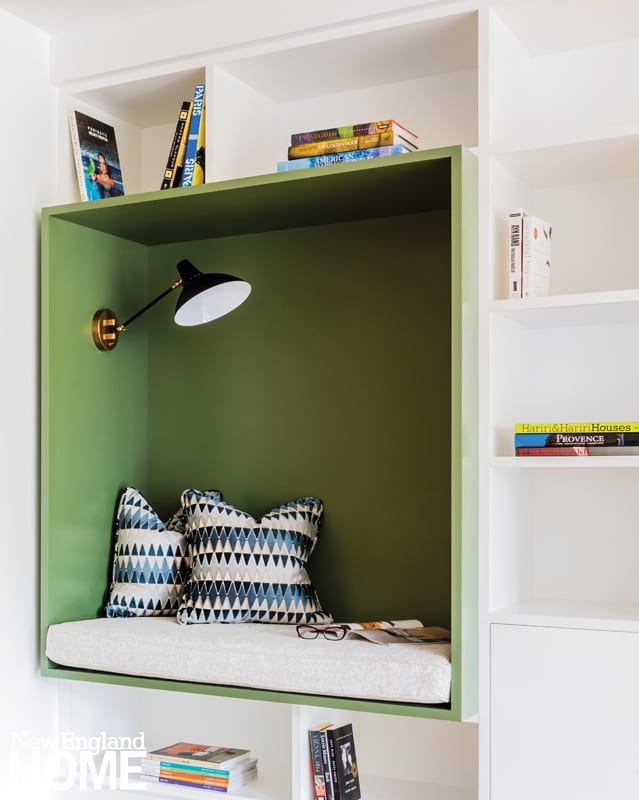 A white wall of built-in shelves with a large green shelf that has been turned into a reading nook complete with a reading light, accent pillows and a white cushion