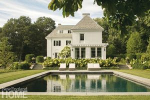 The back of a white house with a swimming pool in front of the house. There are white hydrangeas behind the pool.